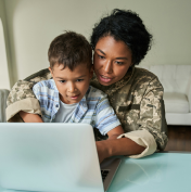 An adult in military uniform helps a child with a laptop at a table in a bright room.